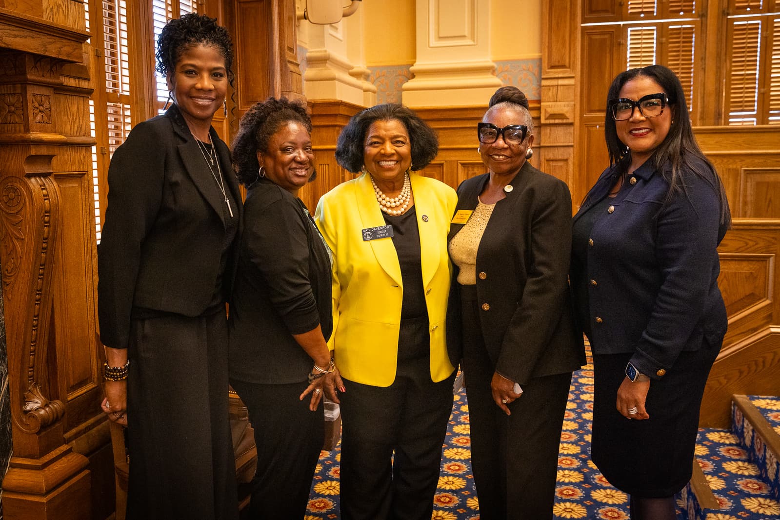 Five professional women pose in a wood-paneled room, one wearing a bright yellow blazer.
