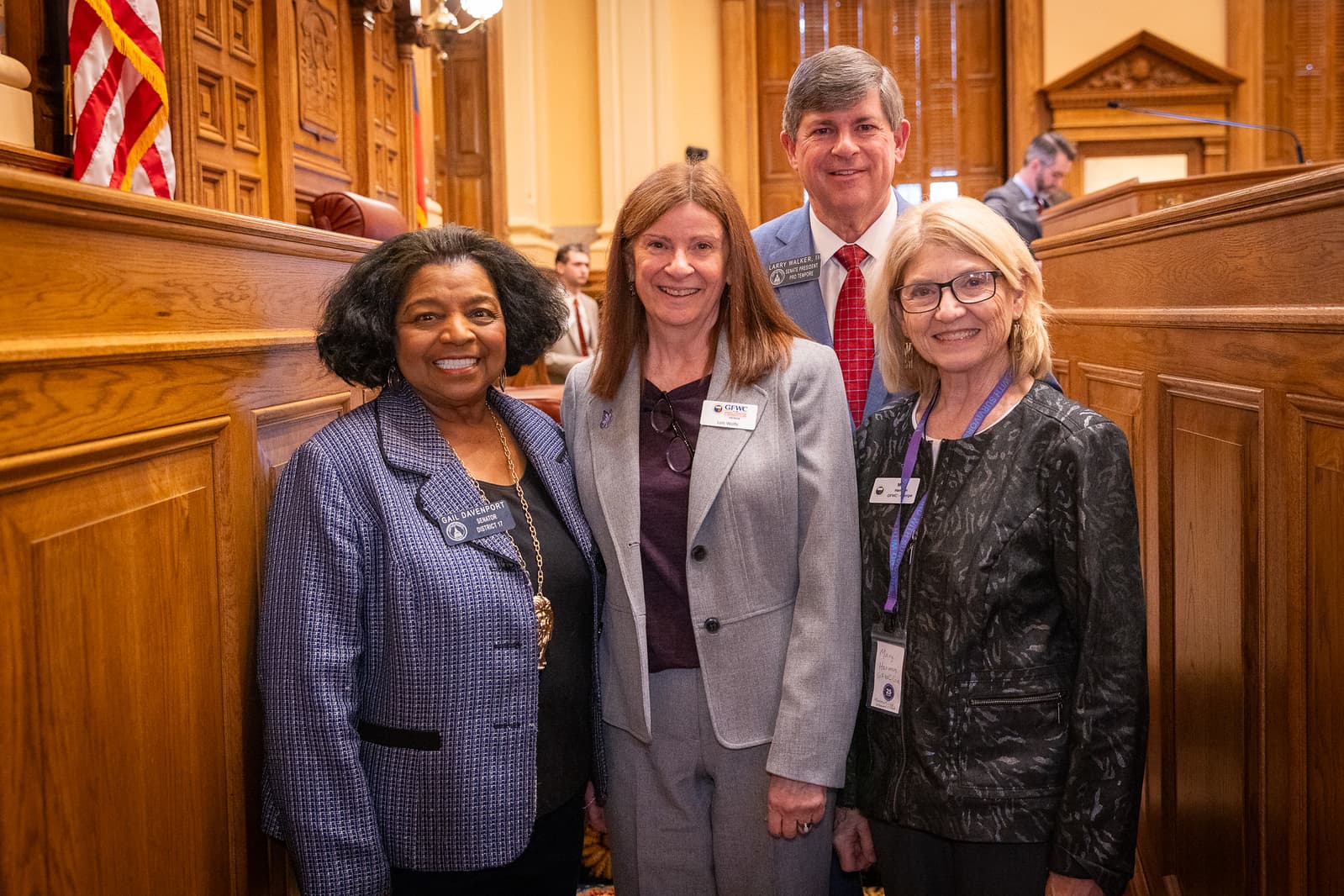 Four smiling professionals in business attire stand together in a formal, wood-paneled legislative room.