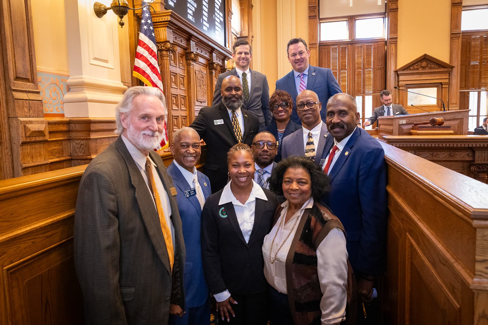 Diverse group of professionals posing for a photo in a formal, wood-paneled legislative chamber.
