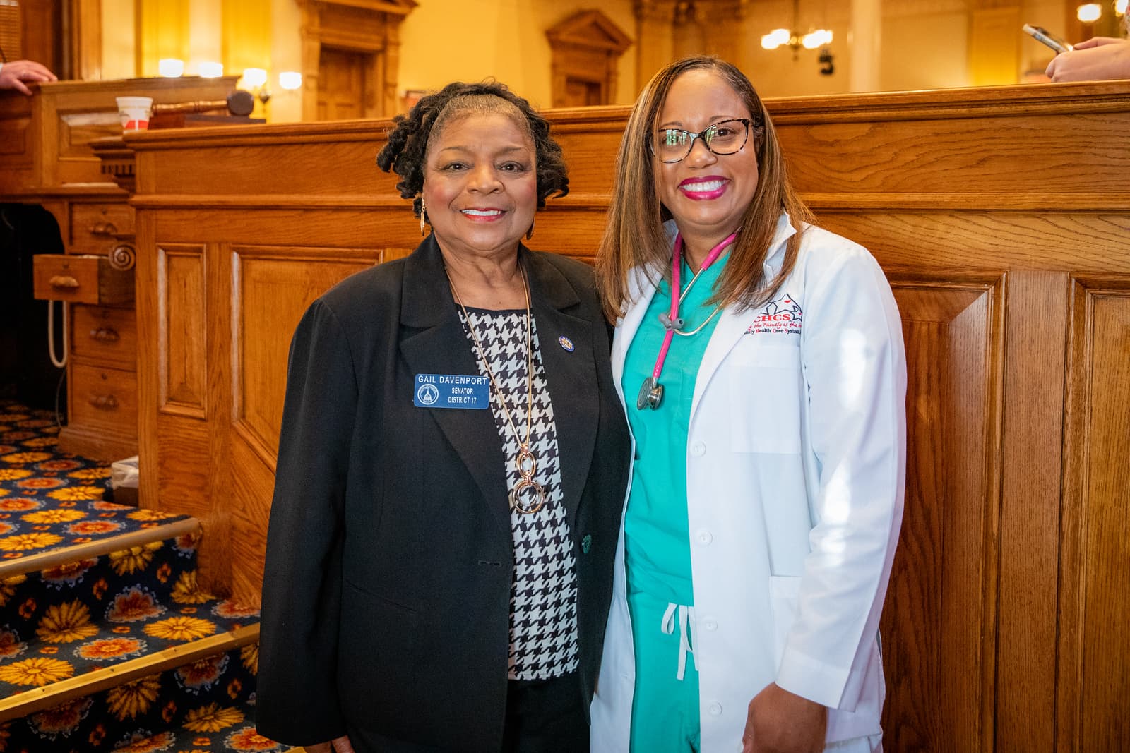 Senator Gail Davenport and a healthcare professional in a white lab coat and stethoscope smile.