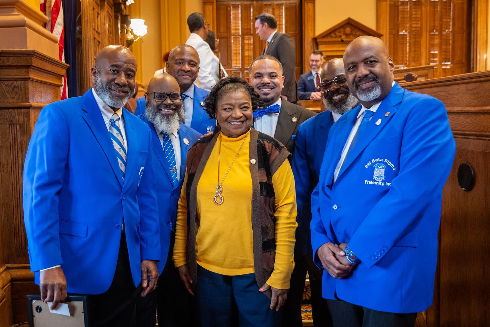 Smiling men in blue Phi Beta Sigma blazers pose with a woman in yellow.