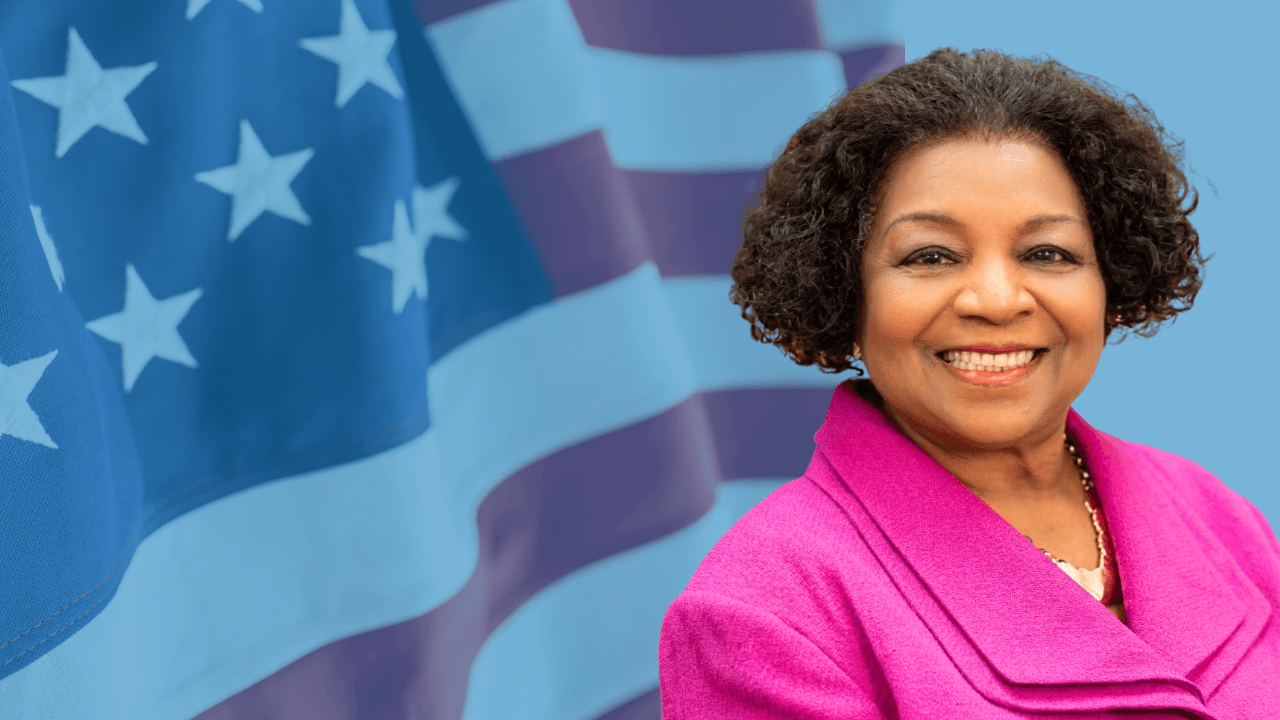 Smiling Black woman in a pink jacket against a blue-toned American flag background.